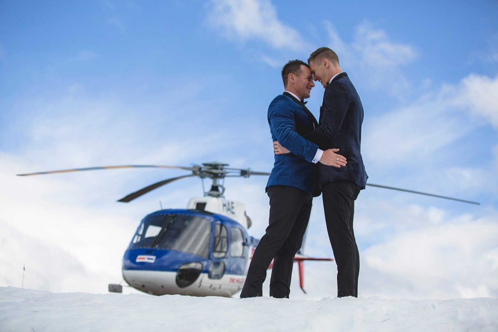 Two groom's hugging on snow with helicopter in background