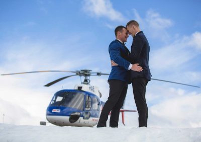 Two groom's hugging on snow with helicopter in background