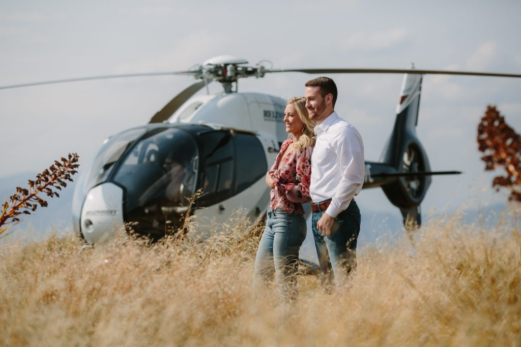 couple standing together in field in front of helicopter